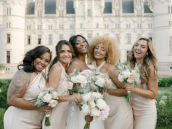 Bridesmaid group photo with bride with bridesmaids in mixed satin dresses holding white and blush rose bouquets before a castle facade