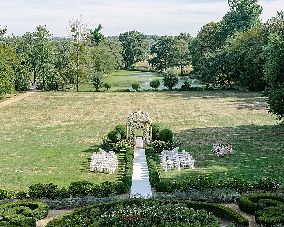 Outdoor ceremony setup with garden wedding ceremony seating, a white aisle runner between hedges leading to a floral arch by a pond