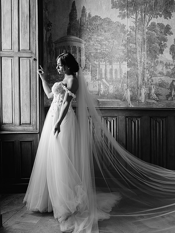 Bridal portrait in black and white of a bride in a long veil, off-the-shoulder lace bodice and tulle skirt, lit by window light.