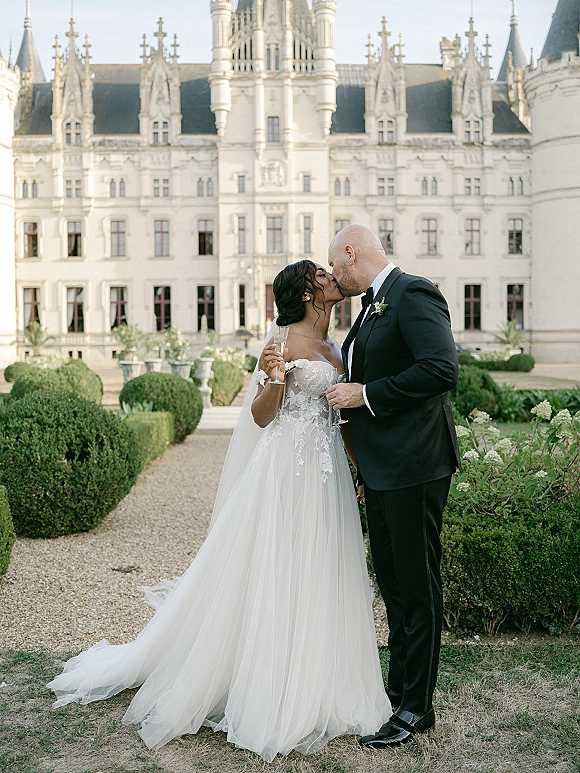 Wedding kiss portrait of bride and groom kissing, bride holding a champagne flute in veil and lace dress before a castle garden path