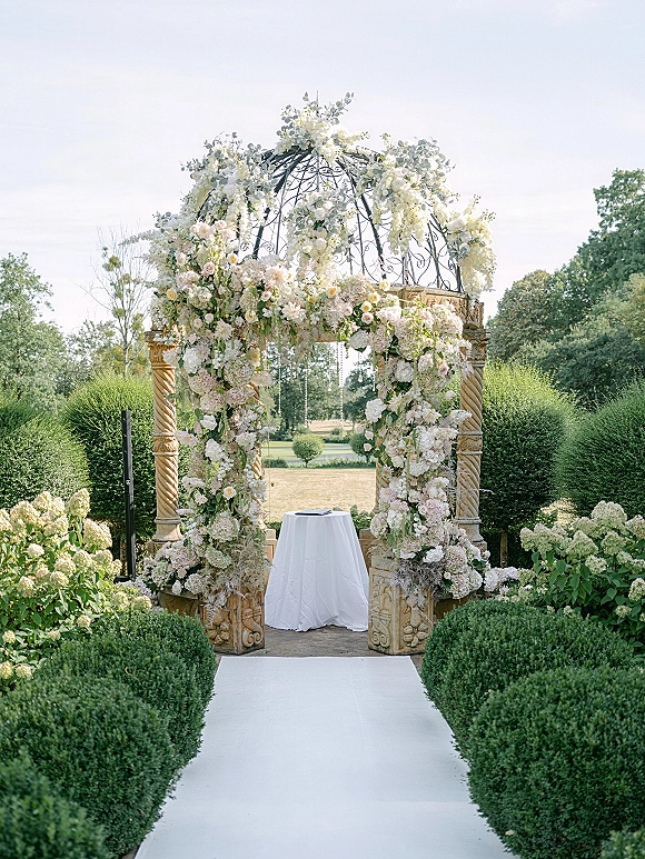 Wedding ceremony altar with a floral wedding arch on a wrought iron gazebo frame, crystal strands, and white aisle runner in a manicured garden