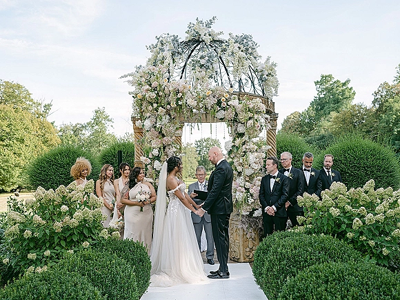 Wedding vows at an outdoor wedding ceremony as bride and groom hold hands under a floral arch with wedding party on a garden lawn