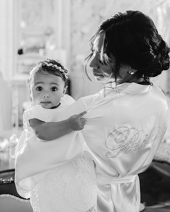 Bride with baby holding her in a lace dress, wearing a monogram bridal robe, looking over her shoulder in soft window light indoors