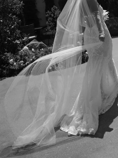 Wedding dress with long wedding veil flowing behind a lace mermaid gown, shown on a sunlit outdoor walkway with garden shrubs