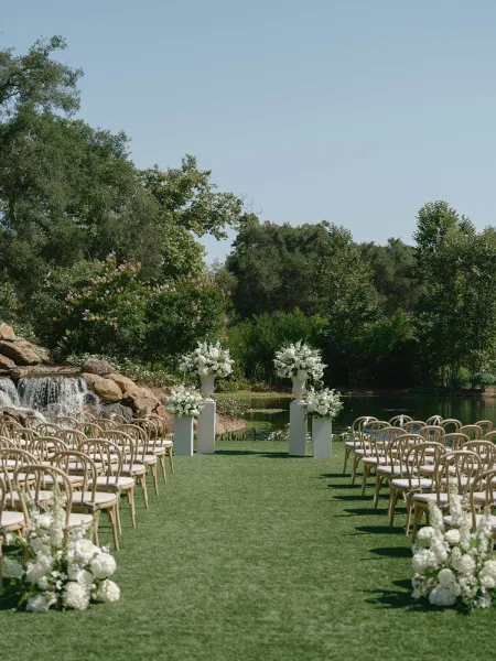 Ceremony aisle design with white floral arrangements on pedestal plinths and urn vases, wooden chairs on a lakefront lawn by a waterfall