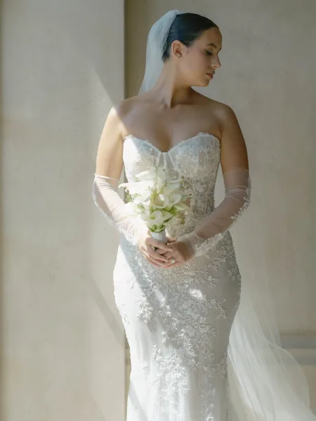 Bridal portrait of a bride in a strapless lace wedding dress with cathedral veil, holding a white calla lily bouquet in soft indoor light