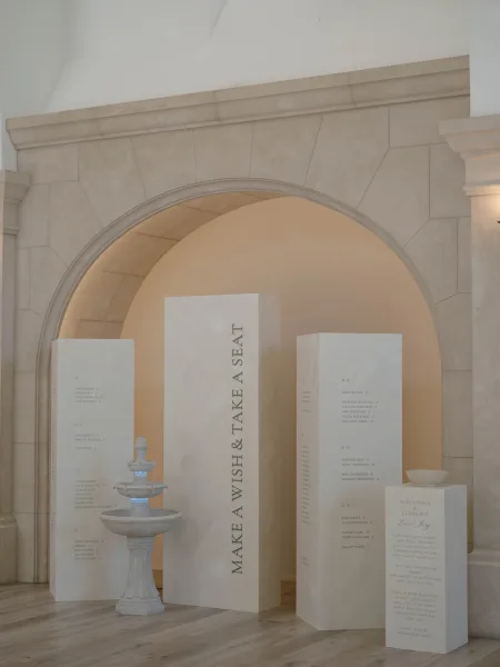 Wedding seating chart with minimalist seating chart pillars beside a stone fountain and welcome sign, set under a stone archway on wood floors