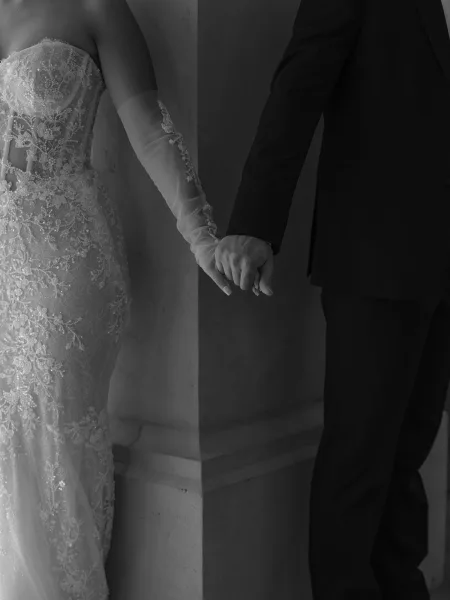 Couple holding hands in a wedding hand holding photo, showing bride’s long gloves and lace beaded gown beside groom’s black suit by stone column