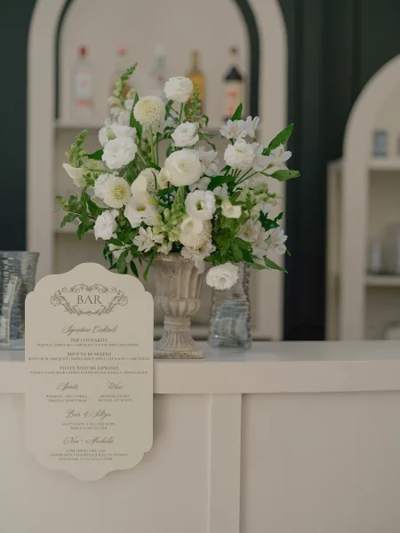 Wedding bar menu and signature cocktail menu card on a white bar counter with ivory scalloped border, floral urn, jars, and liquor bottles behind