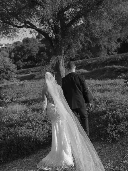 Bride and groom walking away down a dirt path, her long veil trailing behind, beside a tree on a wildflower hillside landscape