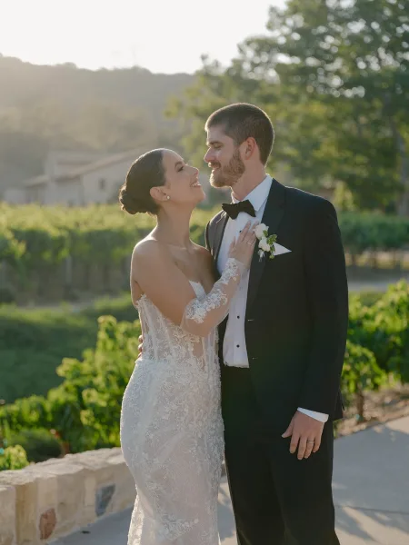 Couple portrait of bride and groom smiling, her hand on his chest, in golden sunset light on a vineyard terrace with stone wall
