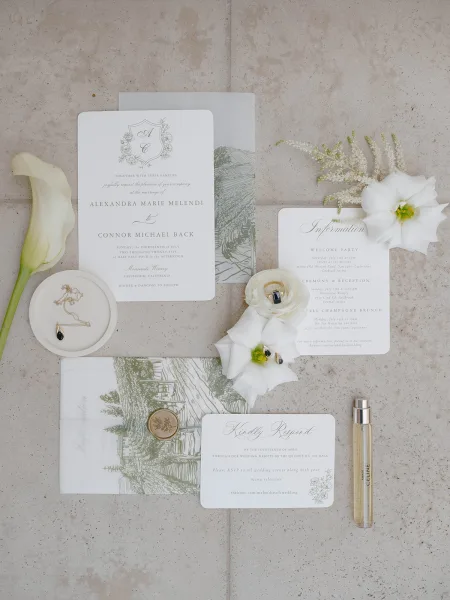 Invitation suite wedding invitations flat lay with wax seal, vellum overlay, envelopes, calla lily, necklace and perfume on stone surface