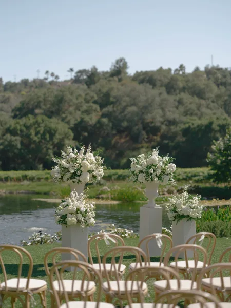 Ceremony setup for an outdoor wedding ceremony with white floral arrangements on plinths, wooden bistro chairs, and pondside lawn views