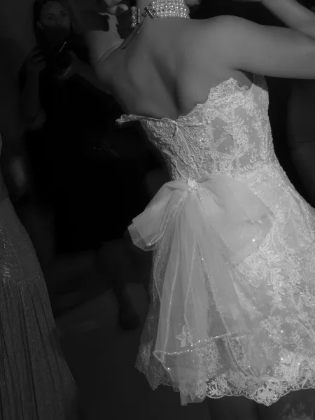Wedding dress close-up of a strapless lace wedding dress with beaded corset bodice and tulle bow, pearl necklace amid dim reception crowd