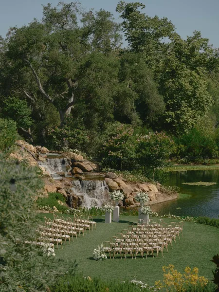 Ceremony setup with outdoor wedding ceremony chairs arranged in a semicircle, white florals lining a curved aisle by pond and waterfall