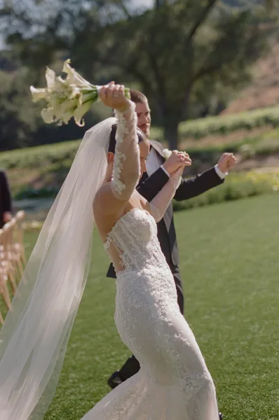 Recessional moment as bride and groom walk down the aisle, just married celebration with bouquet raised on a sunny lawn with trees and hillside backdrop