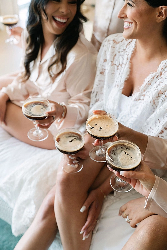 Bridesmaid getting ready in bridal party robes, holding a coupe glass espresso martini in soft window light indoors