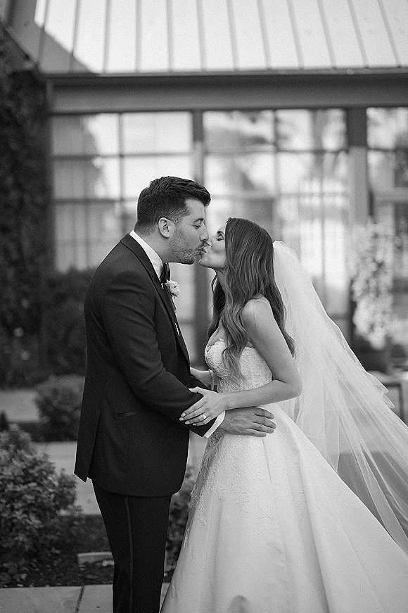 Wedding kiss portrait of bride and groom kissing in side profile, lace dress and long veil, groom in tuxedo by a glass greenhouse walkway
