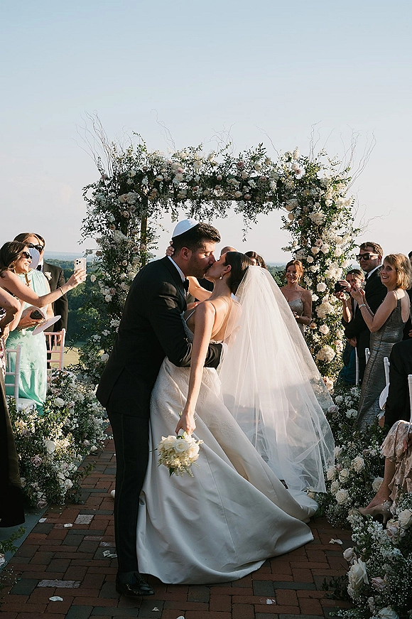 Wedding kiss as bride and groom embrace under a white floral arch with greenery, bouquet in hand, guests watching on brick aisle outdoors