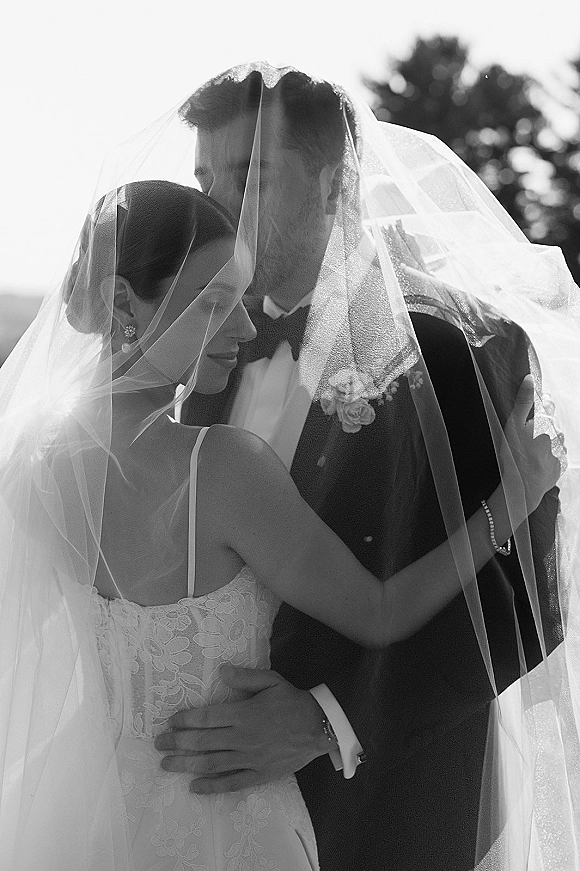 Couple portrait in a black and white wedding portrait style, groom kissing bride’s forehead under her veil, trees and bright sky behind