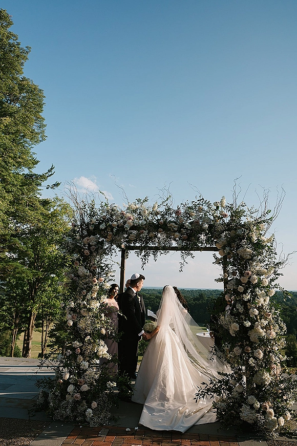 Ceremony moment at an outdoor wedding ceremony as bride and groom hold hands under a rose-and-greenery chuppah on a stone patio