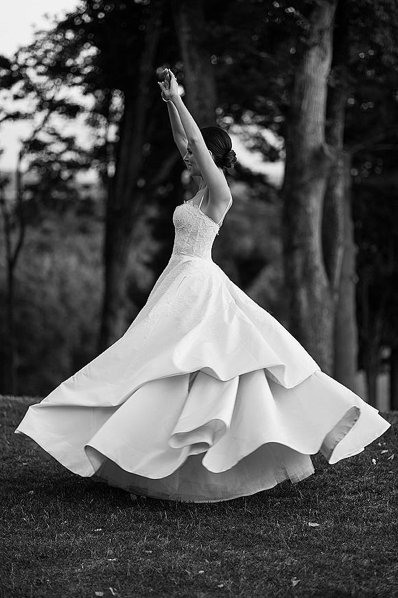 Bridal portrait in black and white of a bride twirling her strapless lace ball gown, arms raised, with an updo in a park lawn by trees