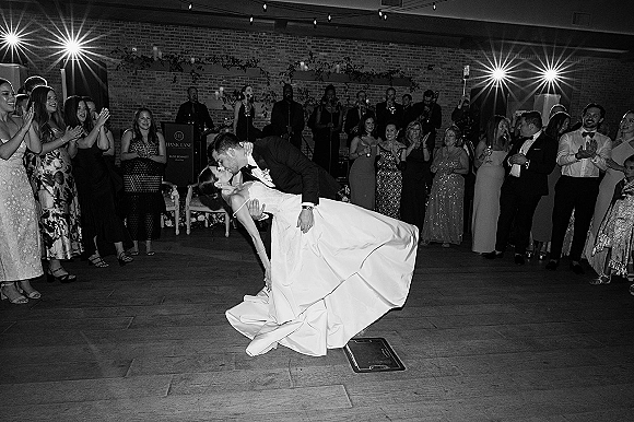 First dance dip as bride and groom kiss on the dance floor, candles and string lights glowing, live band before brick wall and guests