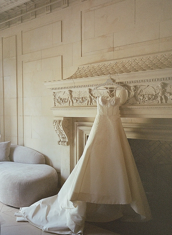 Wedding dress with lace wedding dress details hanging on a hanger, long train draped before an ornate fireplace mantel in a paneled room