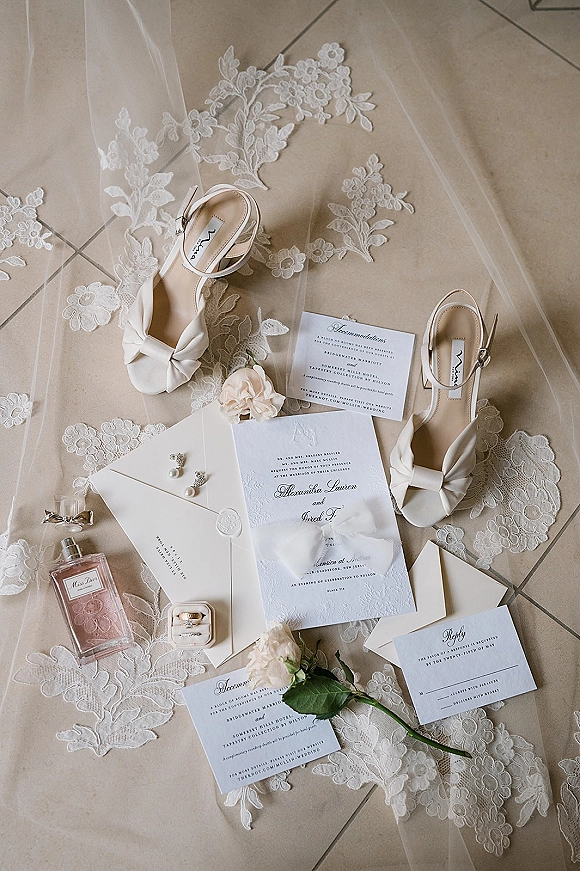 Invitation suite wedding invitation flat lay with envelopes, RSVP and details cards, wax seal, ribbon, rings, heels, and pearl earrings on tile floor