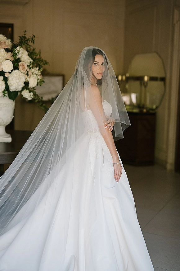 Bridal portrait of a long veil bride in a strapless gown, looking over her shoulder in a neutral indoor hallway beside florals