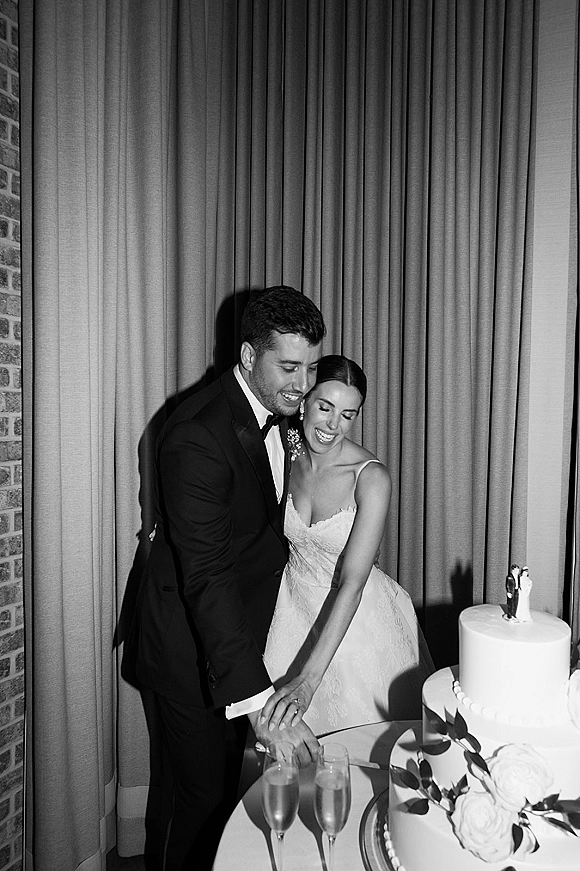 Cake cutting moment as bride in strapless gown and groom in black tuxedo slice a two-tier white cake on a table by brick wall