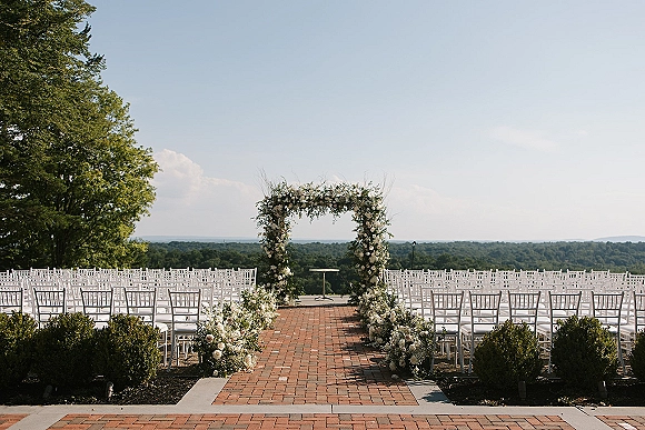 Ceremony setup with an outdoor wedding ceremony floral arch, white flowers and greenery lining a brick patio aisle with chairs and hills beyond