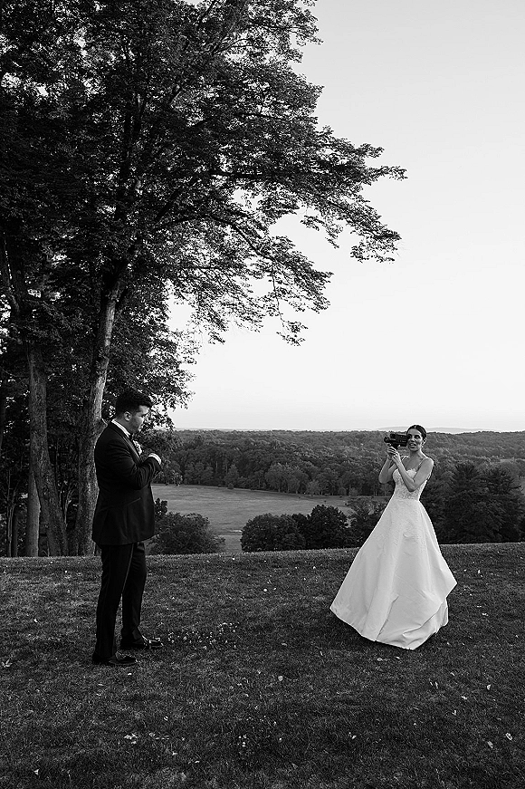 Wedding first look as bride films groom with a camcorder, her ballgown and his tuxedo under a tree on a scenic lawn with hills