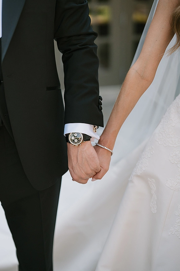 Couple holding hands, bride and groom hands with black tuxedo sleeve, wedding watch and bracelet, walking an outdoor greenery walkway