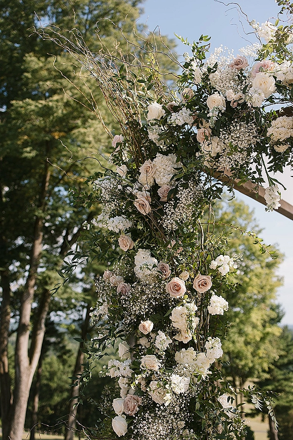 Wedding ceremony arch with asymmetrical roses and hydrangea accents on a wooden frame, set on a lawn with trees and blue sky