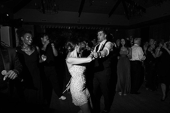 Wedding dance floor couple dancing reception as the bride in a strapless beaded mini dress spins with her groom under chandeliers, guests cheering