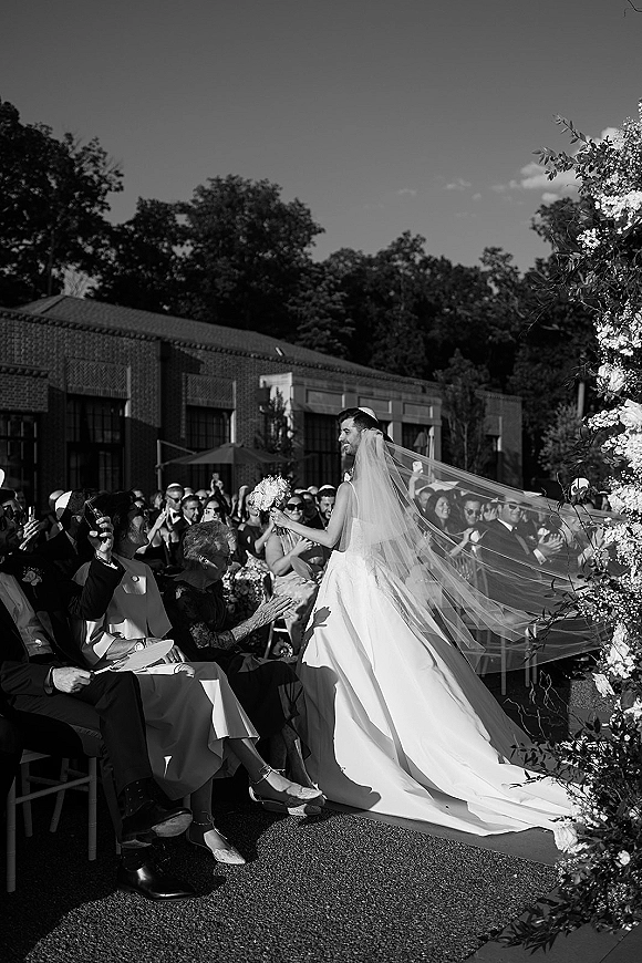 Wedding processional with bride walking down aisle in a strapless gown, bouquet in hand, long cathedral veil trailing past seated guests outdoors