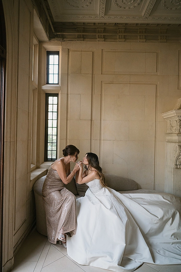 Bridal portrait of a bride in a strapless wedding dress with a long train, seated by tall windows in a stone-walled suite