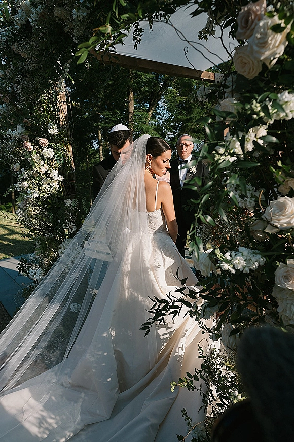 Wedding ceremony moment as bride in long veil and gown stands with groom in tuxedo and kippah under a white rose chuppah in sunlit garden