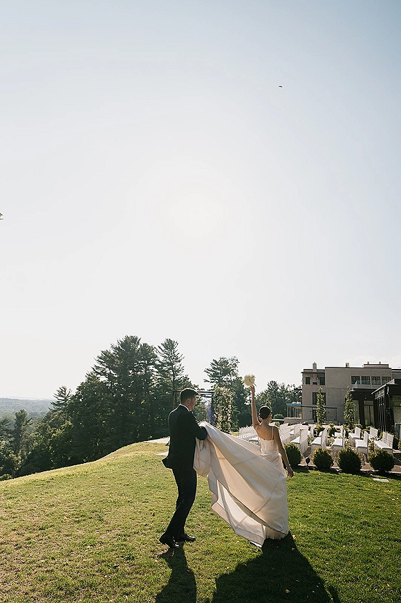 Wedding recessional as bride and groom walk away on a lawn aisle, bride lifts bouquet, groom carries her train, modern venue behind