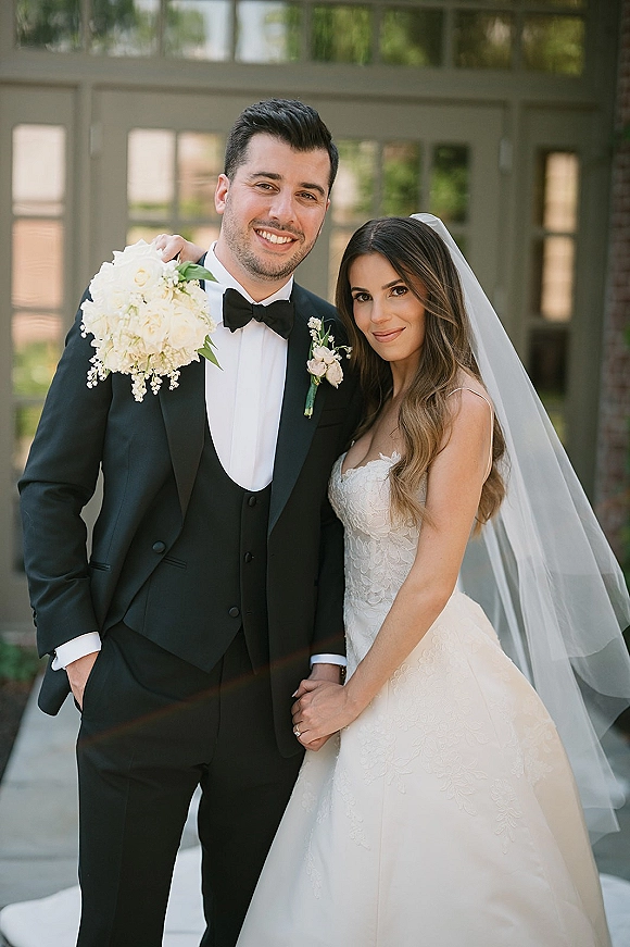 Couple portrait of bride and groom holding hands, her lace dress and veil with white bouquet, beside glass doors and brick wall greenery