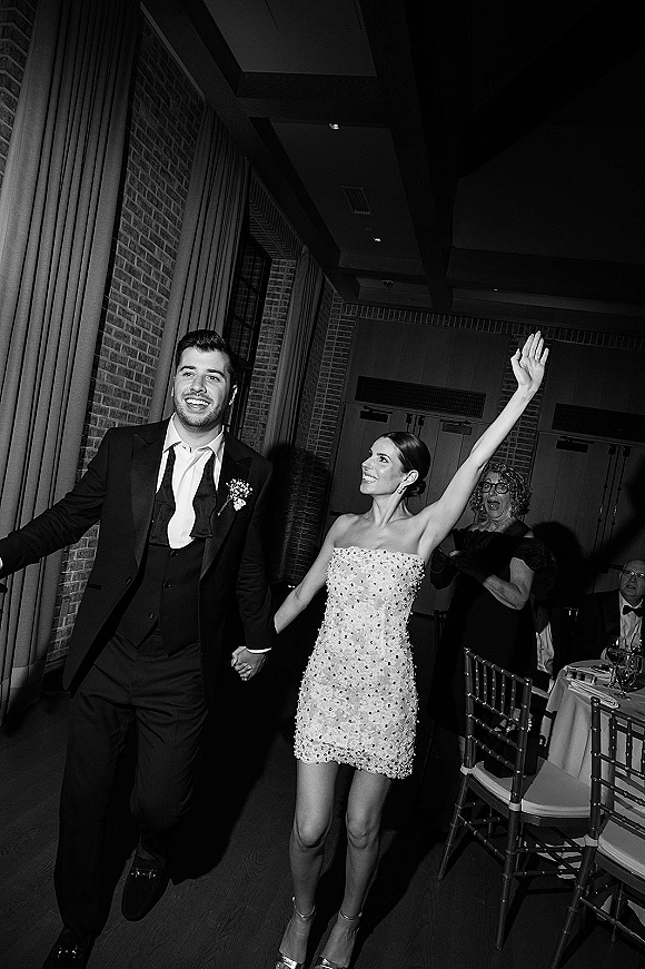 Wedding reception entrance as newlyweds hold hands, bride in strapless mini dress waving beside groom in tuxedo by brick wall doors