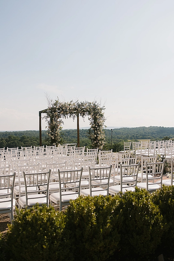 Ceremony setup for an outdoor wedding ceremony with a floral wedding arch, white chiavari chairs, and mountain-view terrace backdrop