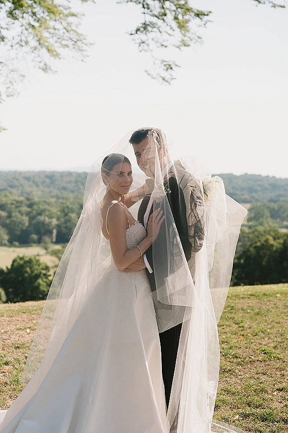 Couple portrait of bride and groom under veil, embracing in a hilltop meadow with trees and distant forested hills under open sky