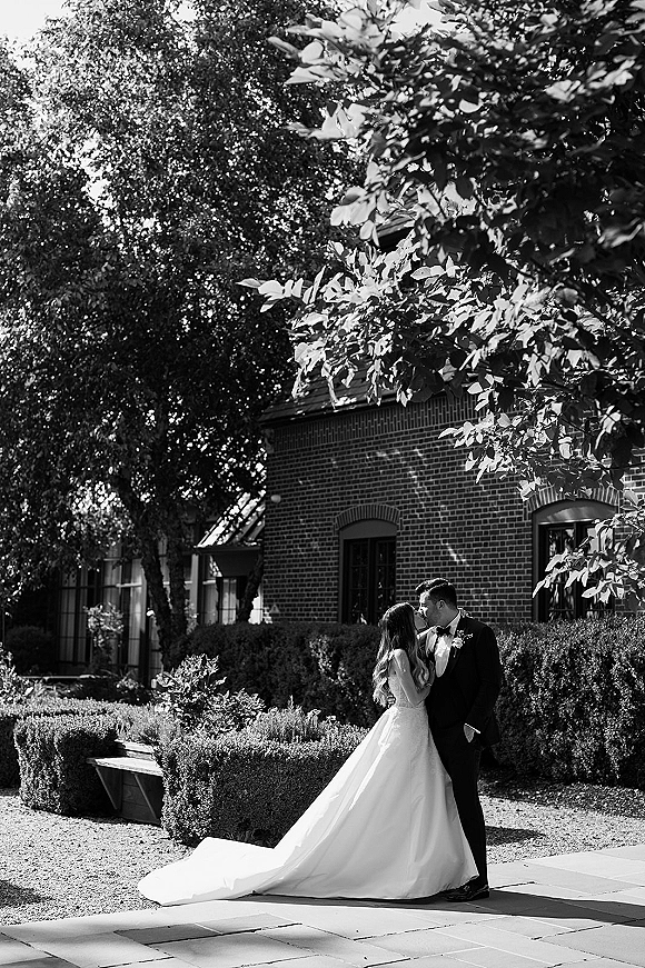 Wedding kiss portrait of bride and groom kissing on a stone walkway, her long train flowing as he wears a bow tie near brick windows