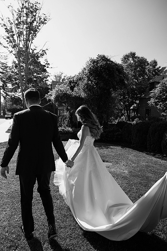 Wedding couple portrait of newlyweds holding hands, walking away on a lawn as the bride’s long train and veil trail behind near trees and a house
