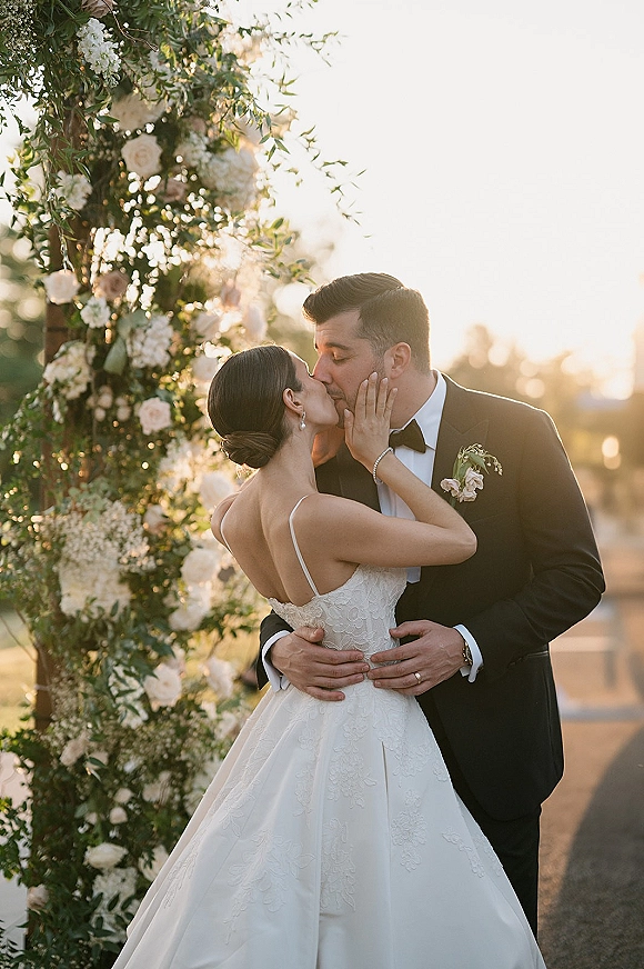 Wedding kiss portrait of bride and groom kissing beneath a white rose and greenery floral arch at sunset, bride’s hand on his face