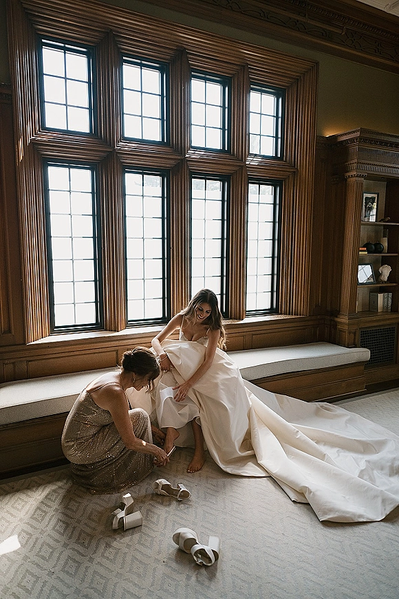 Bride getting ready as a bridesmaid helps with her strapless gown and long train by large windows in a wood-paneled suite