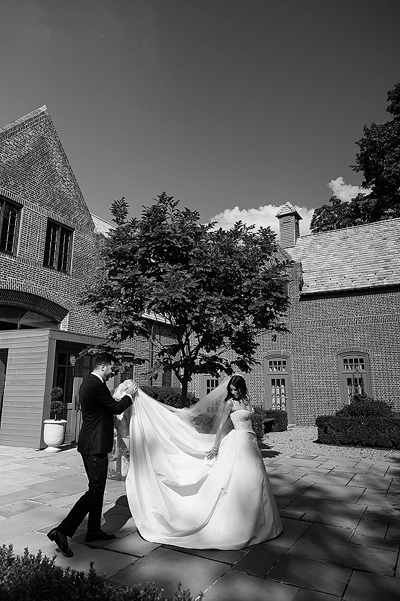 Couple portrait of bride looking over her shoulder as groom holds her long dress train in a brick courtyard, veil flowing behind