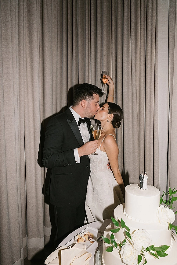 Wedding kiss portrait of bride and groom kissing beside a two-tier white cake with champagne flutes on a curtained table backdrop